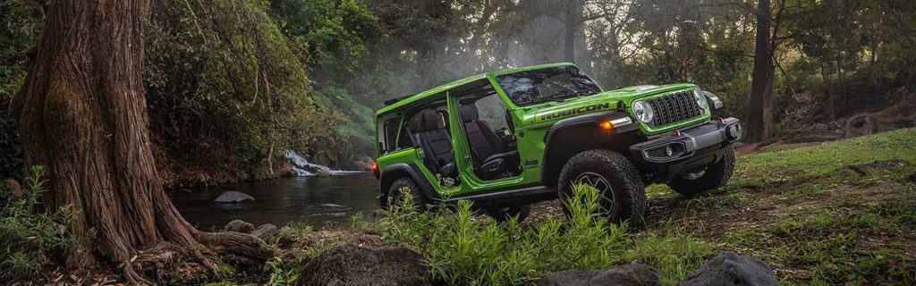 A green Jeep Wrangler Rubicon parked by a river in a forested area. 
